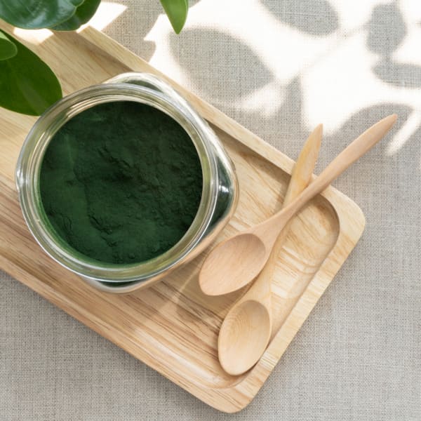 a glass jar full of spirulina powder sitting on a wooden board beside two wooden spoons