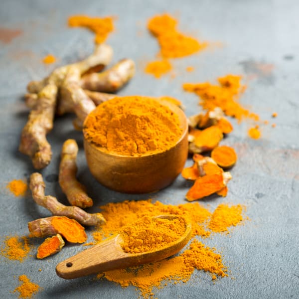 a small wooden bowl filled with turmeric powder beside a wooden spoonful of powder alng with pieces of fresh turmeric root
