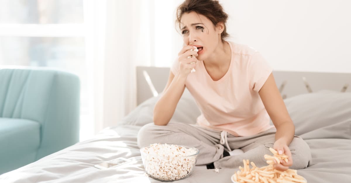 woman sitting on bed crying and eating unhealthy food