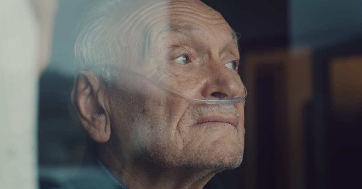 An elderly thoughtful man wearing an oxygen tube is looking through a window