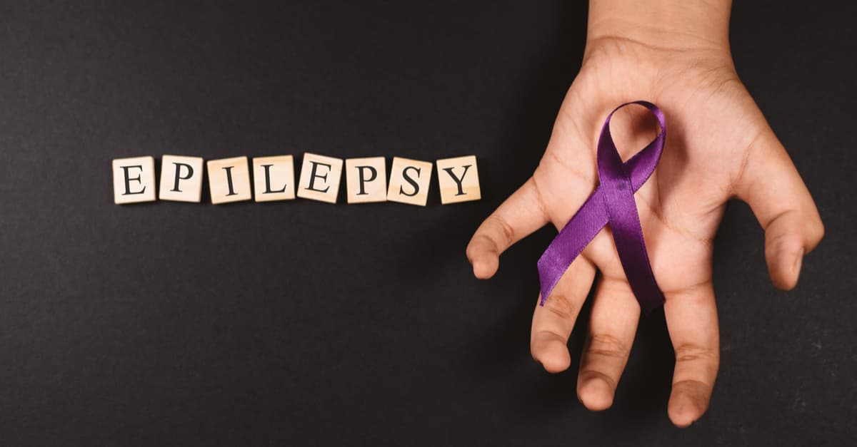 Purple ribbon on cramped hand andthe word EPILEPSY against black background.
